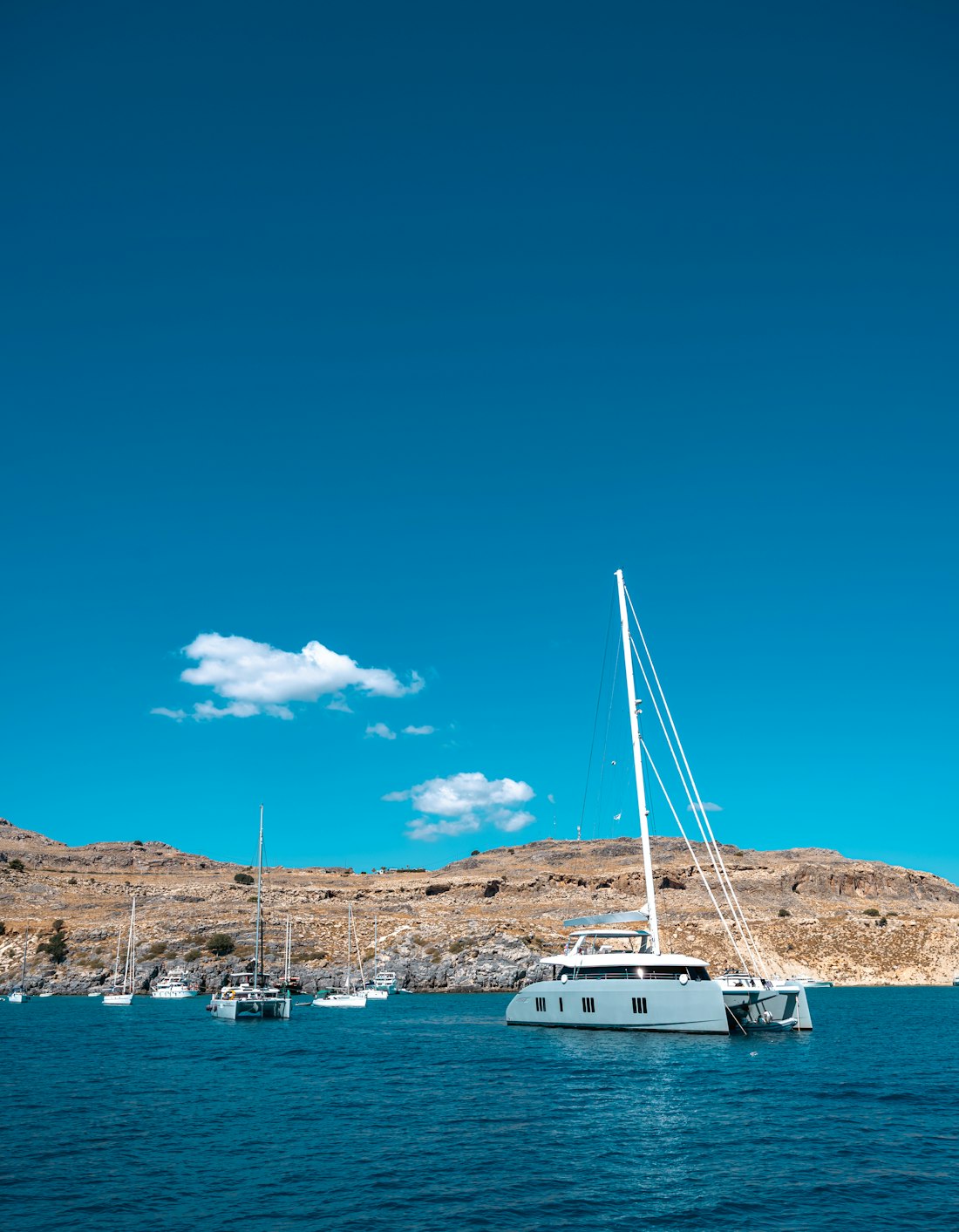 A sailing catamaran anchored by arid rocky shores in clear turquoise water